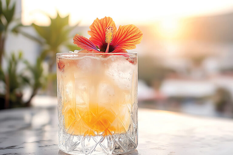 Glass with ice and orange cocktail decorated with hibiscus flower on marble table, with blurred cityscape and sunset in background mindful drinking